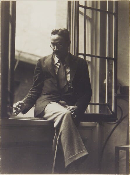black and white photo of a man sitting cross-legged on a windowsill. He's wearing a jacket, tie and slacks, and is smoking a cigarette.
