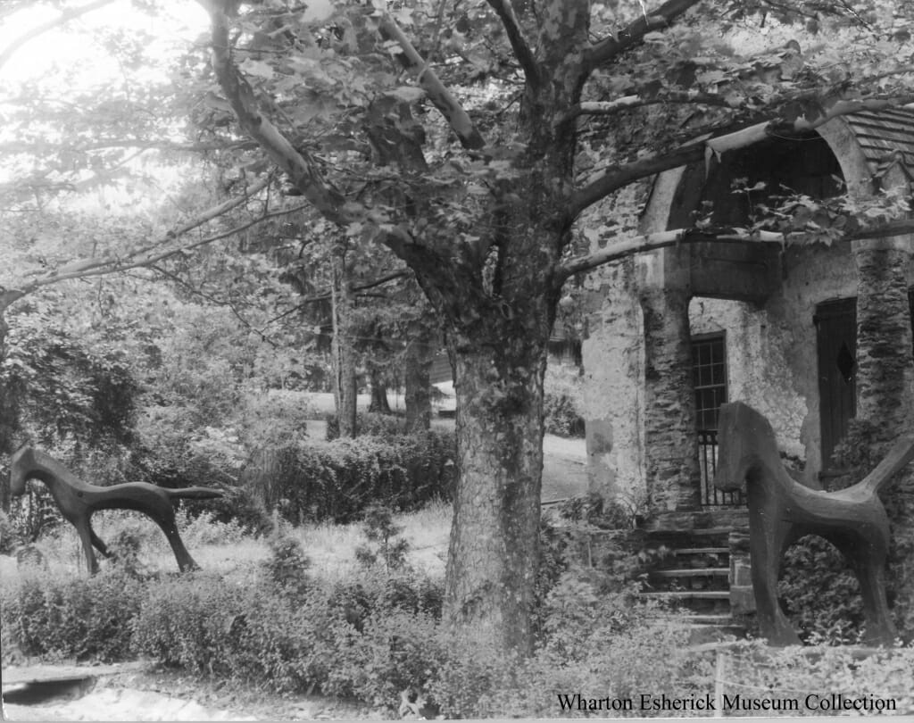 Black and white photo with large tree in the center and two large wood horse sculptures on either side, all in front of stone building with stone archway over front door.