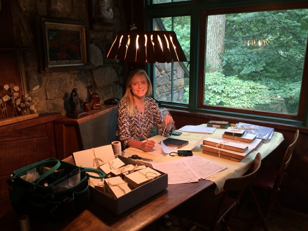 Young woman sits at table surrounded by woodcut blocks and paperwork.