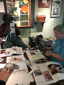 Two women seated around a table full of open books work on paper chair models