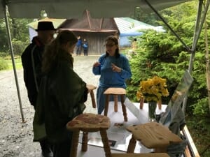 Three people standing outside under a tent next to a table with short three-legged stools on it.