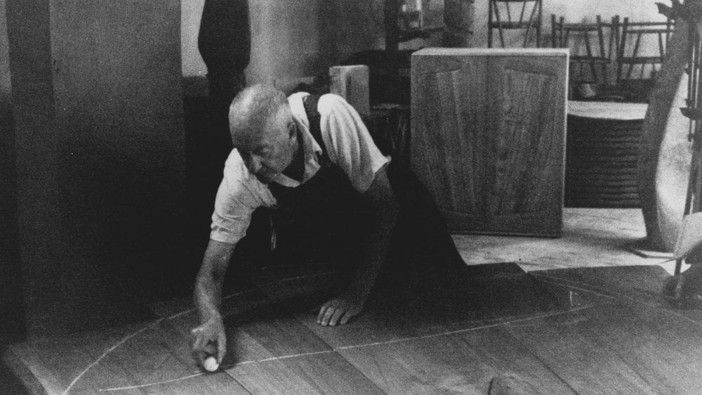 Black and white photo of older mandrawing a chalk line across wood boards in a workshop.