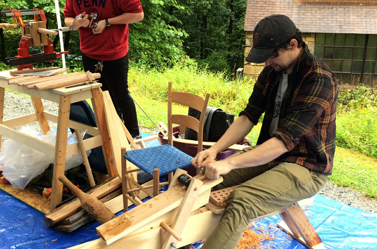 Young man using drawknife to shape a chair leg. Chair in background.