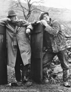 two men leaning on the gate of a stone wall, with farmland around them.