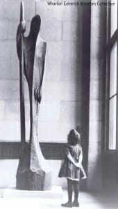 a young girl looks up at a 12 foot tall wood carved sculpture of a man leaning on a staff