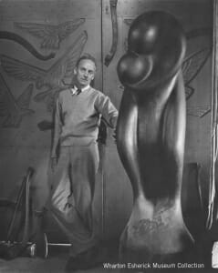 man (Wharton) in sweater and slacks leans against carved wood loading doors with large wood sculpture by his side. sculpture is rounded abstration of two people in an embrace.