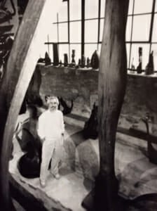 man standing among many tall wood sculptures, windosill of small wood sculptures behind him