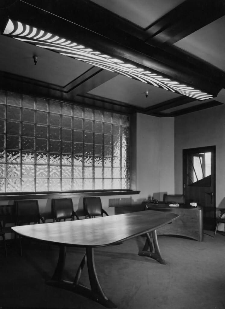 organic curving boardroom table with glassblock window behind and ceiling light with curving patterned grille over long rectangular light