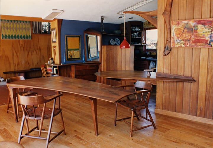 dining area of home with wood walls with blue accents. Rectangular but curving dining room table surround by captain's chairs in the center of the space with cantilevered breakfast counter and entrance to kitchen in the background
