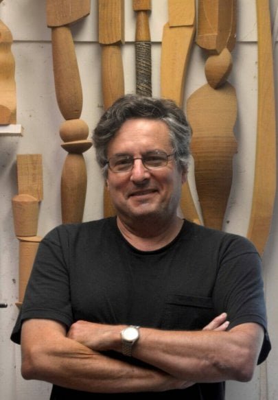 man in black t-shirt with arms folded across chest is smiling. Turned wooden forms hang on a wall behind him.