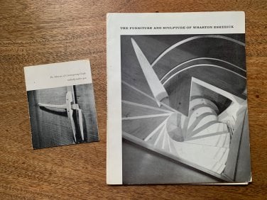 A black and white exhibition catalog and a smaller notecard-sized invitation lay on a solid wood desktop. The catalog features the title 'The Furniture and Sculpture of Wharton Esherick' and a view looking down at a spiraling staircase made for the Curtis Bok house. The invitation includes a black and white photo of a slender wooden door latch with the words 'The Museum of Contemporary Crafts cordially invites you' written above.