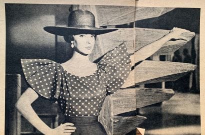 black and white photo of woman from the waist up, wearing polka dot blouse with pronounced, slightly ruffled sleeves, and a wide brimmed hat. she has her right hand on her hip and her left hand on the cantilevered step of a wooden spiral staircase behind her