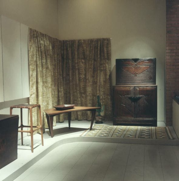 view of gallery showing several pieces of wood furniture wrapped around two wall of the room. From left to right stand a four legged stool with a woven leather seat, an asymmetrical coffee table with a darker wood bowl on top and a carved wood cabinet and desk unit. These is also a greenish-tan curtain hanging in the corner of the room, and a woven rug on the floor in front of the desk unit.