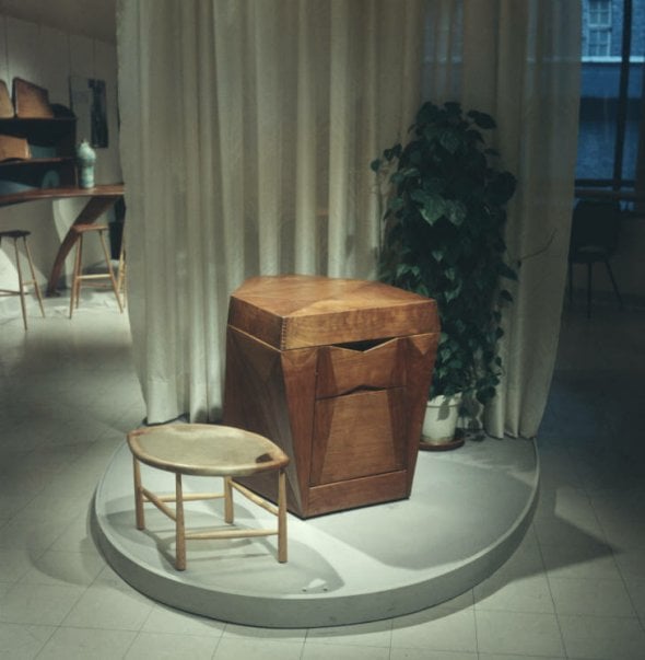 ovoid plinth in the center of a gallery with a short wood stool with an almond shaped seat made of rawhide next to a wood phonograph cabinet. The cabinet is a boxy form, but has faceted asymmetrical planes. there is a soft white curtain behind them and a houseplant. far behind the curtain we can see a couple stools and other furniture.