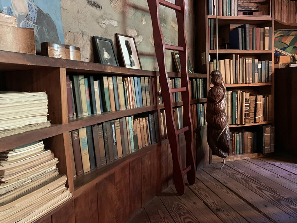 two rows of books to the left lead back to another vertical bookshelf. a red ladder leans in front of the two shelves and a carved wood sculpture hangs from a rope beyond the ladder. small items and photo lean against the wall above the bookshelves and the floor is pine wood boards.