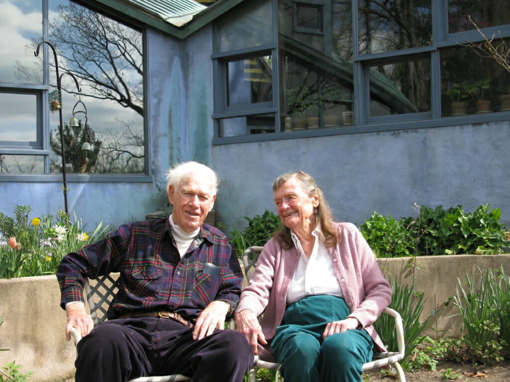 An elderly couple sit outside in front of a blue stucco building with large windows. The man is wearing corduroy slacks, a white turtleneck and a dark plaid shirt and smiling at the camera. The woman is wearing teal pants, and a pink cardigan over a collared shirt. She is turned toward the man and smiling
