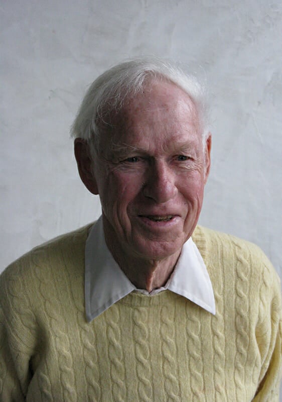 Elderly man with a collared shirt and pale yellow sweater and white hair is smiling and seated in front of a gray wall