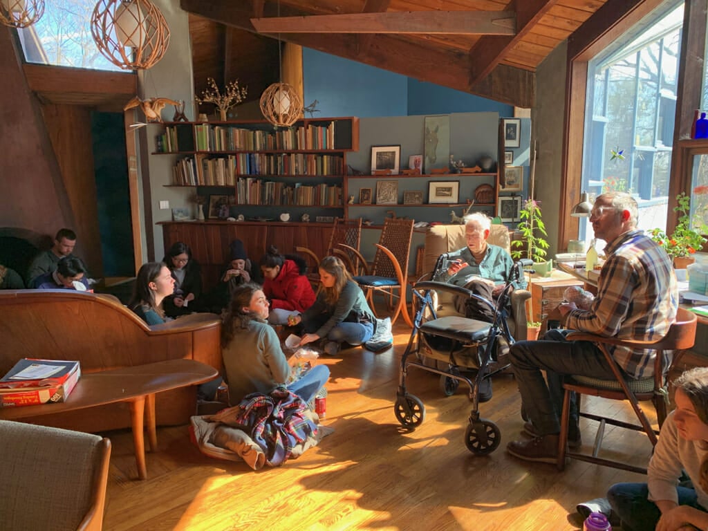 a group of college students sit on the floor in a large light-filled room listening to an older man in an armchair.