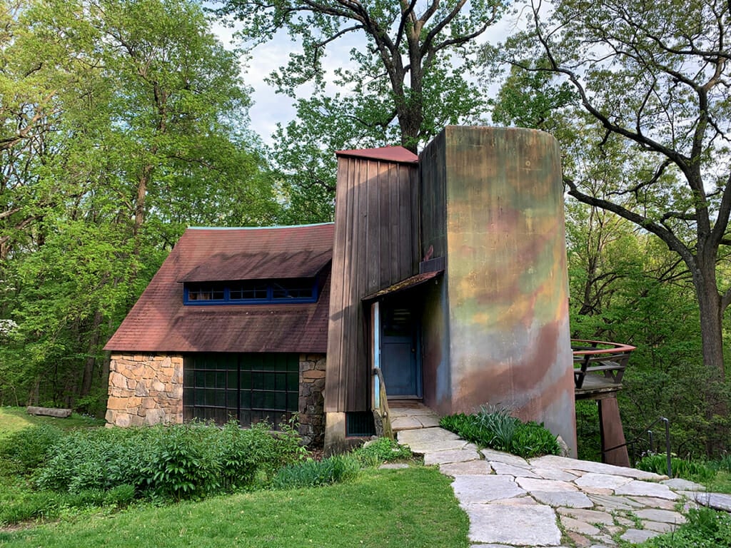 studio building with stone section on the left, tall wooden addition in the middle and colorful stucco on the right, surrounded by green grass with forest in the background