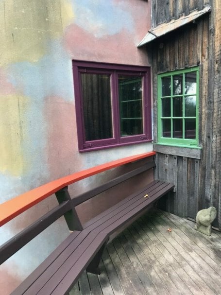 view of kitchen windows from the deck. Red railing and brown bench end in a corner with plum window on the left wall and green window on the right.