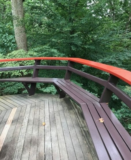 view of deck shows curving red railing and bench with woods in the background
