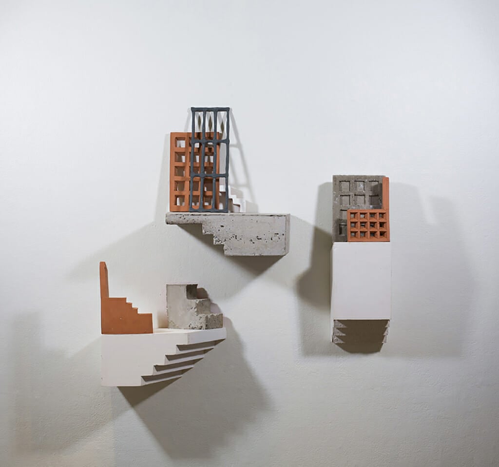 three sculptures on a white wall depict staircase and building like forms