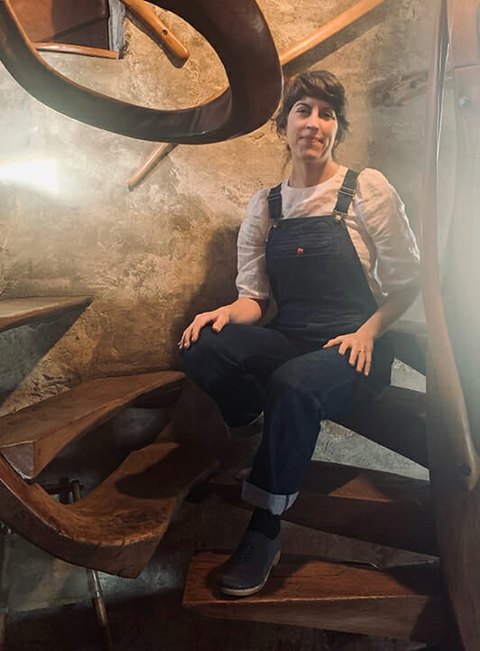 woman in white shirt and overalls sits on wooden spiral steps with beams of light shining on either side of her