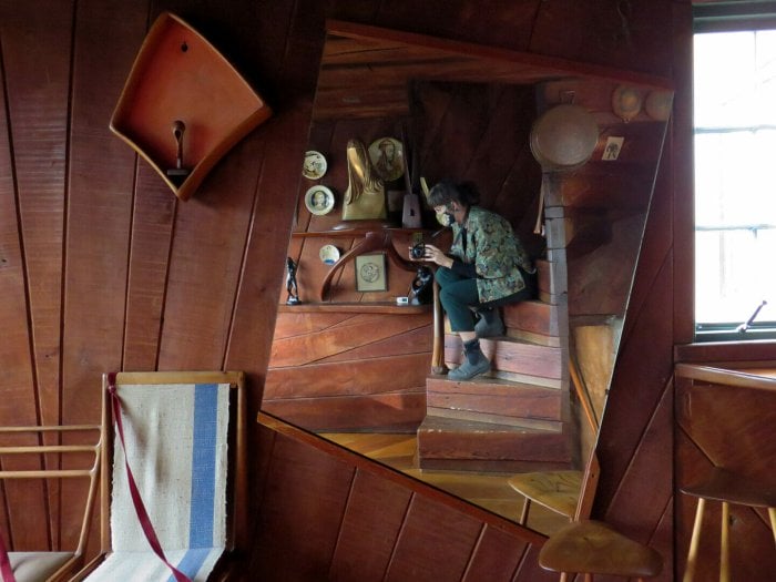 wood board wall with asymmetrical mirror on wall. In the reflection is a woman crouching on a wooden spiral staircase.