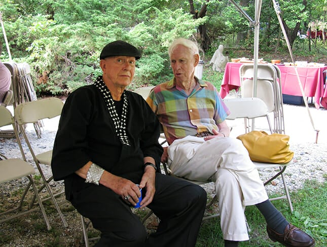 Two older men sit outside on folding chairs. One wears all black, with a black cap, and silver armband. The other wears white slacks and a pastel plaid button-down shirt.