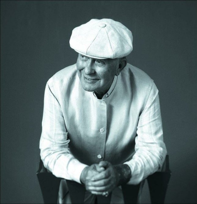 a older man in a white cap and white shirt leans forward, resting on his elbows with his hands held together. He looks off to his right slightly and has a friendly gentle smile.
