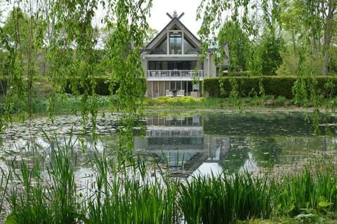 An a-frame house sits across a pond. There are hanging green tree limbs, green grasses, lilypads, and a hedge around the pond.