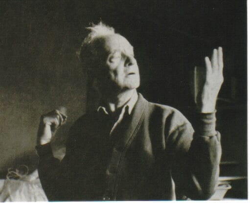 old man wearing shirt and cardigan gestures with one hand up and the other behind his back
