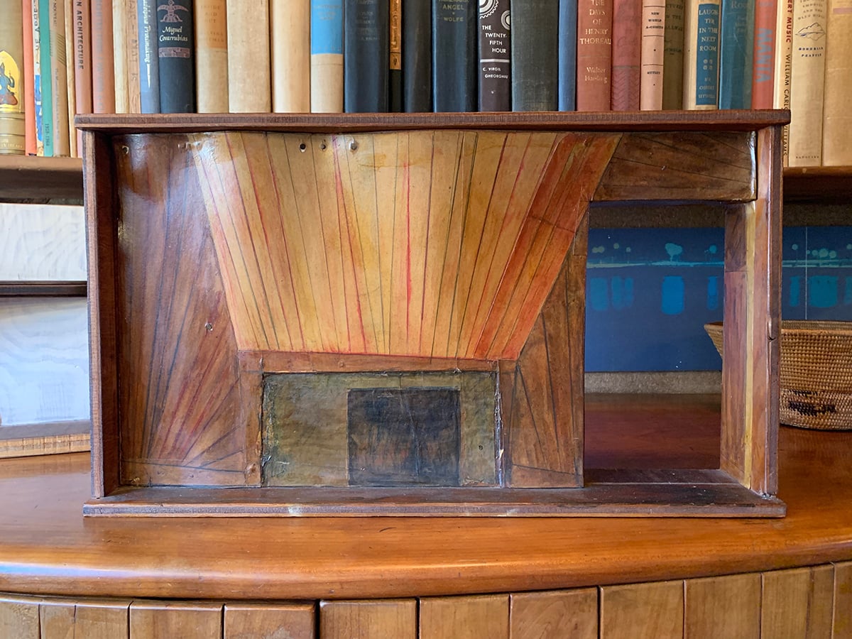 wood and paper model of a fireplace sits on a cabinet in front of a shelf of books