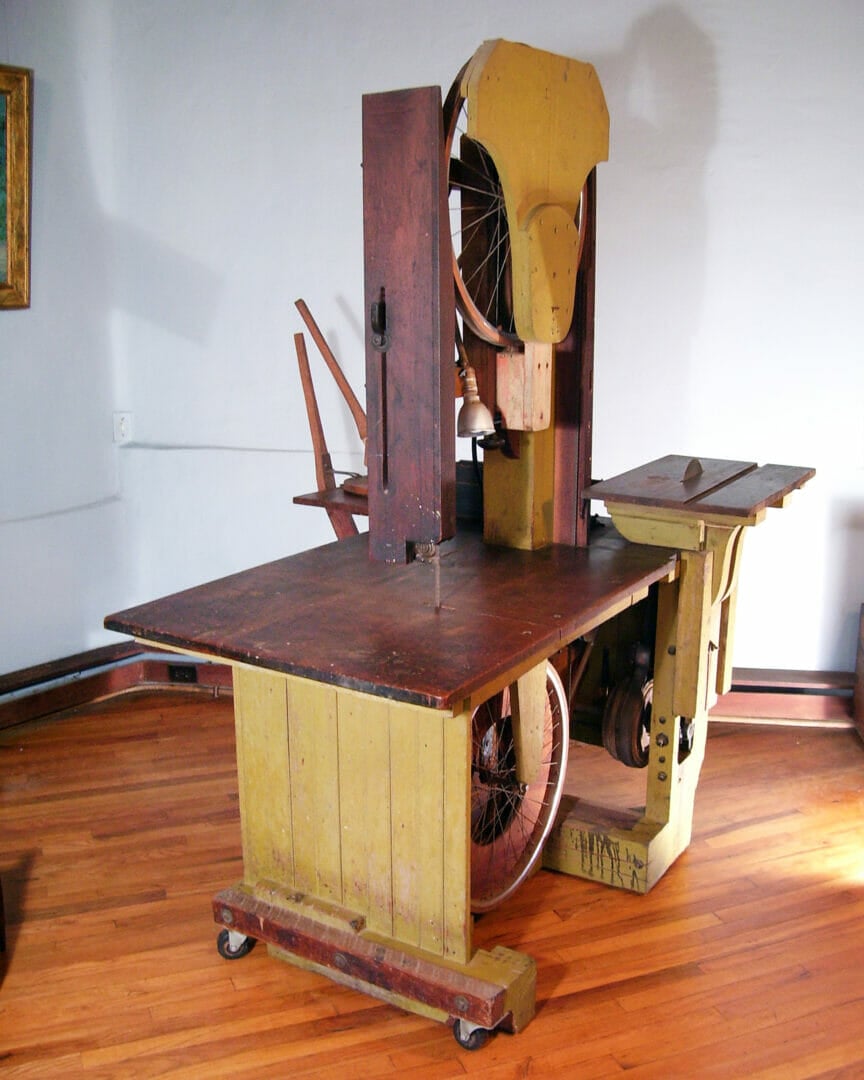 a homemade bandsaw made of wood and bicycle wheels sits in a room with a wood floor and pale lavender walls. The table surface of the bandsaw is a dark wood and the vertical sides of the tool are painted a mustard yellow.