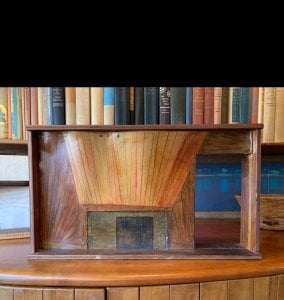 wood and paper model of a fireplace sits on a cabinet in front of a shelf of books