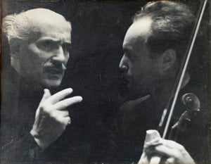 black and white photo shows head and shoulders of two men speaking closely. Man on left is bald with wispy white hair on the sides of his head and has a mustache. He has an expression of serious thought on his face and is gesturing with his right hand. The other man has dark hair and is facing the other man, listening. We can see in the photo he is holding a violin.