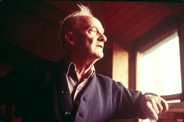 Wharton Esherick when he is older looks thoughtfully out a window as afternoon light illuminates his face. He leans his arm on the windowsill and is wearing a collared shirt and a dark blue cardigan.