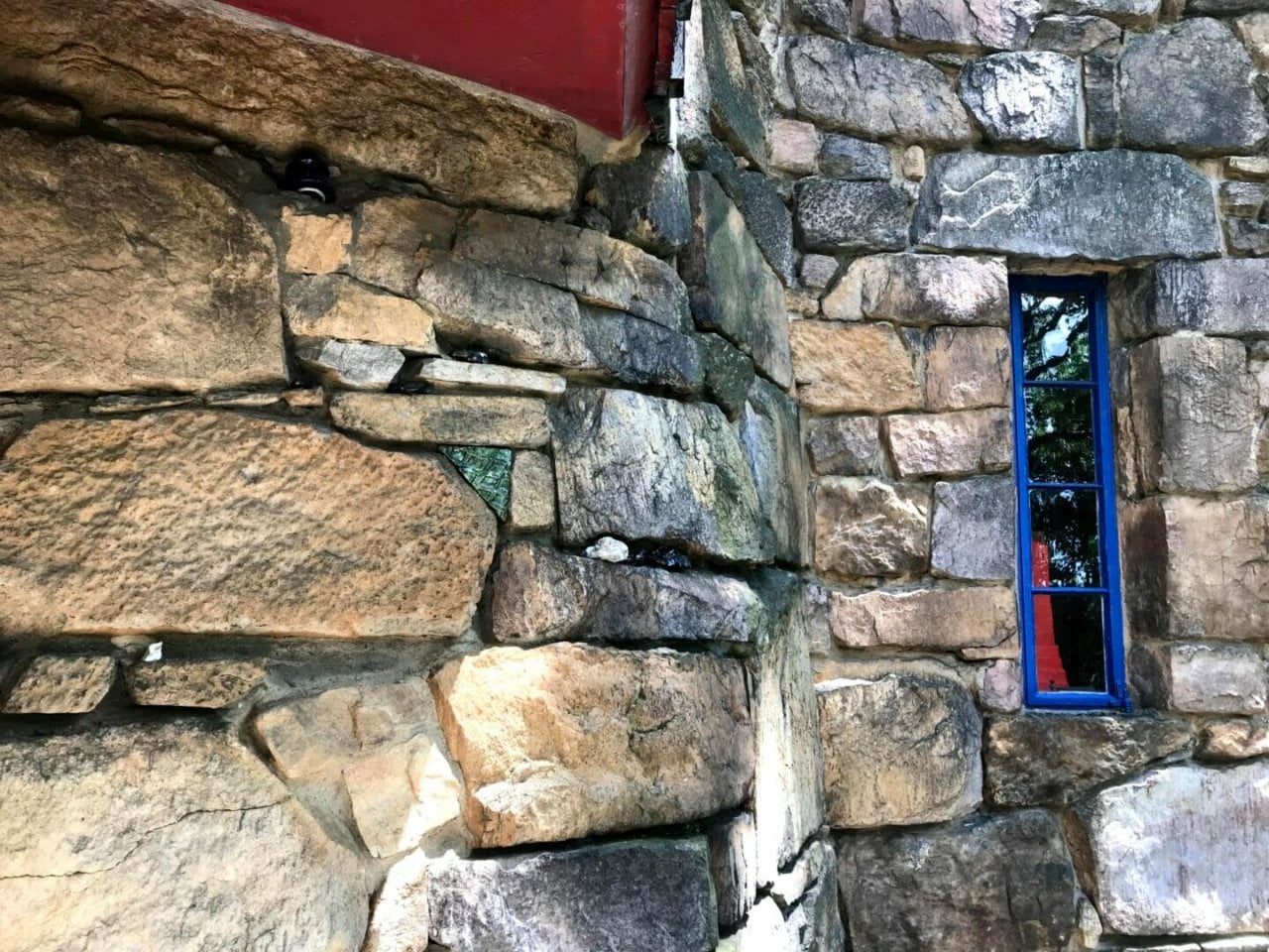 A stone wall with a tall narrow, blue window towards the right. In the window we can see a bright red abstract sculpture.