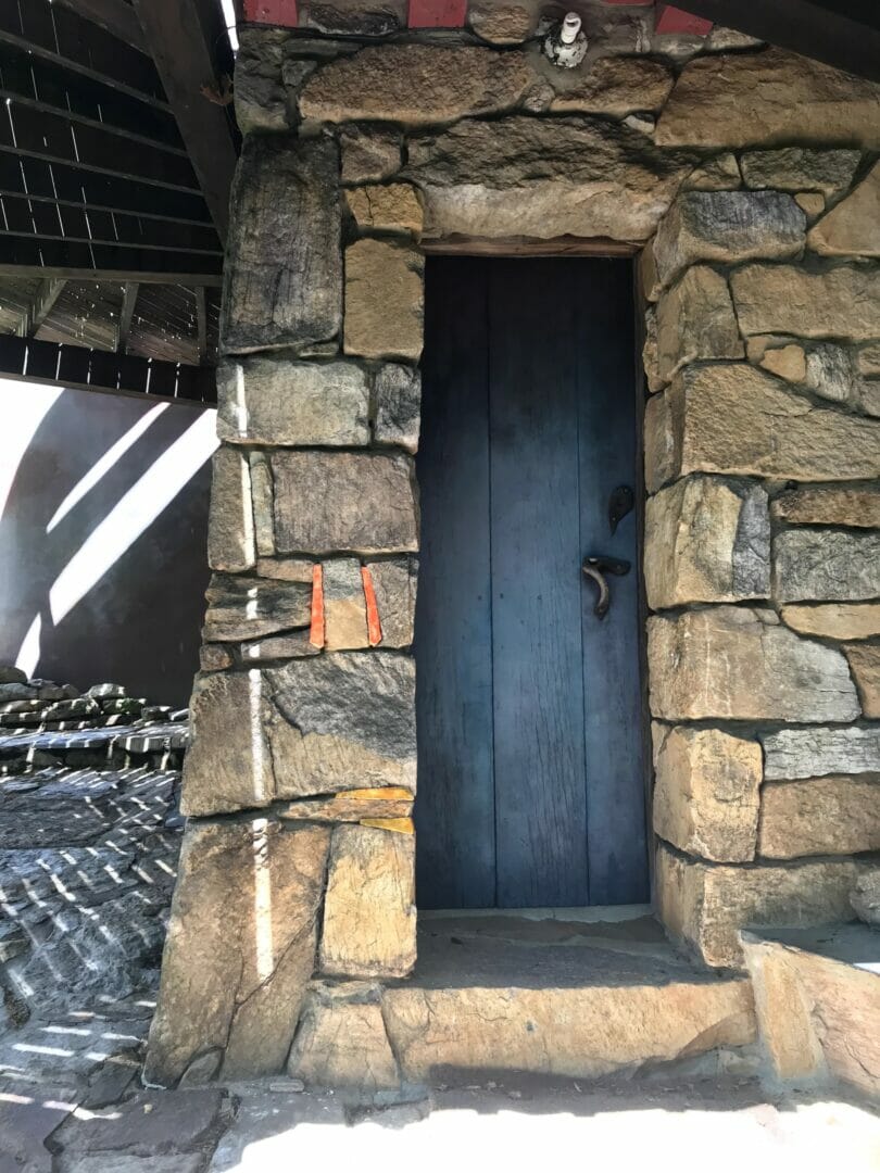 A blue door set in a stone wall is under a deck. Orange and yellow ceramic pieces fir the the cracks in the stone wall