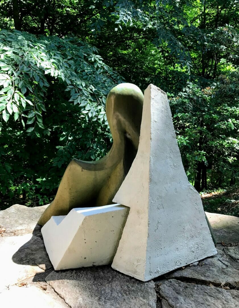 a bronze pelican sculpture is partially obscured by angular white forms. They sit on a stone patio with green woods in the background.