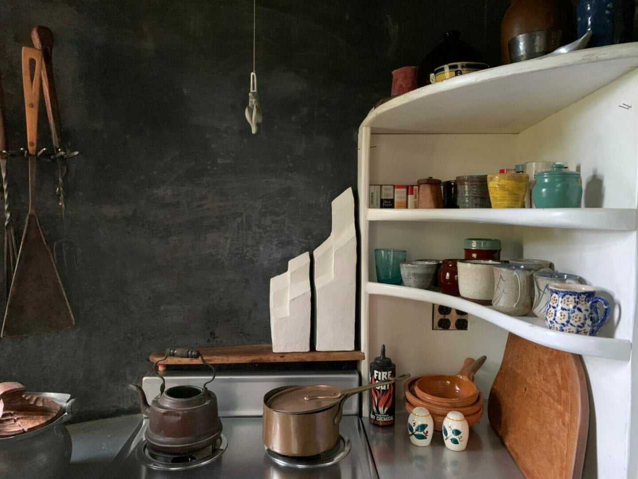 two stepped white sculptural forms sit on a shelf behind a kitchen stove. White shelves with kitchen items extend to the right.